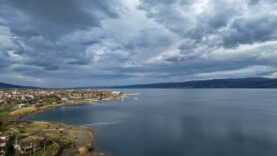 Göl kenarında, bulutlu gökyüzü altında sakin ve geniş su yüzeyi.