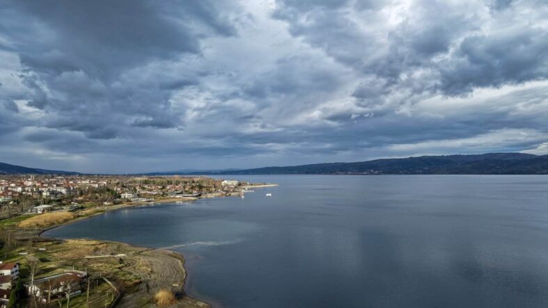 Göl kenarında, bulutlu gökyüzü altında sakin ve geniş su yüzeyi.